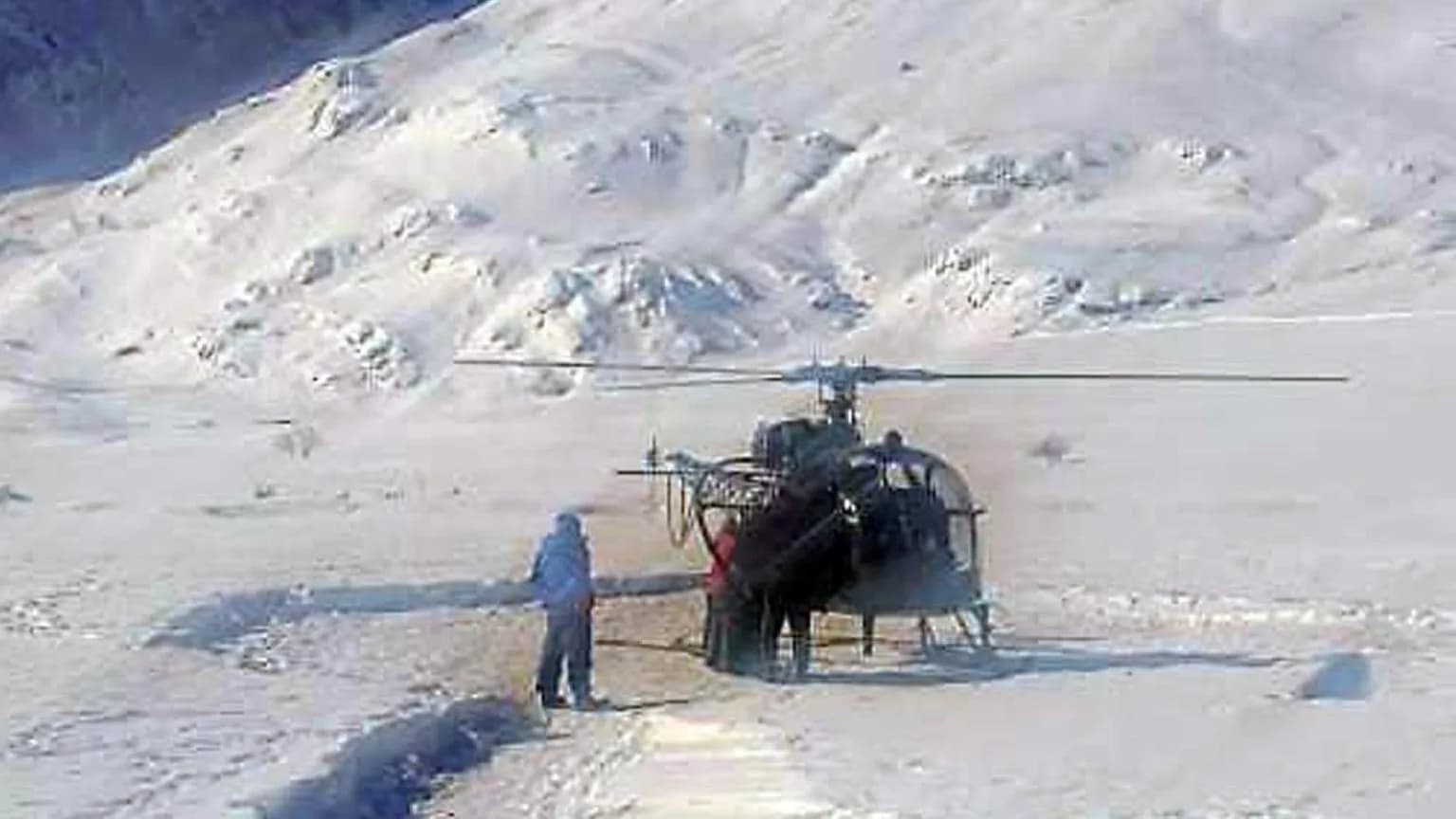 A helicopter lands on a snowy mountainside with a person standing nearby.