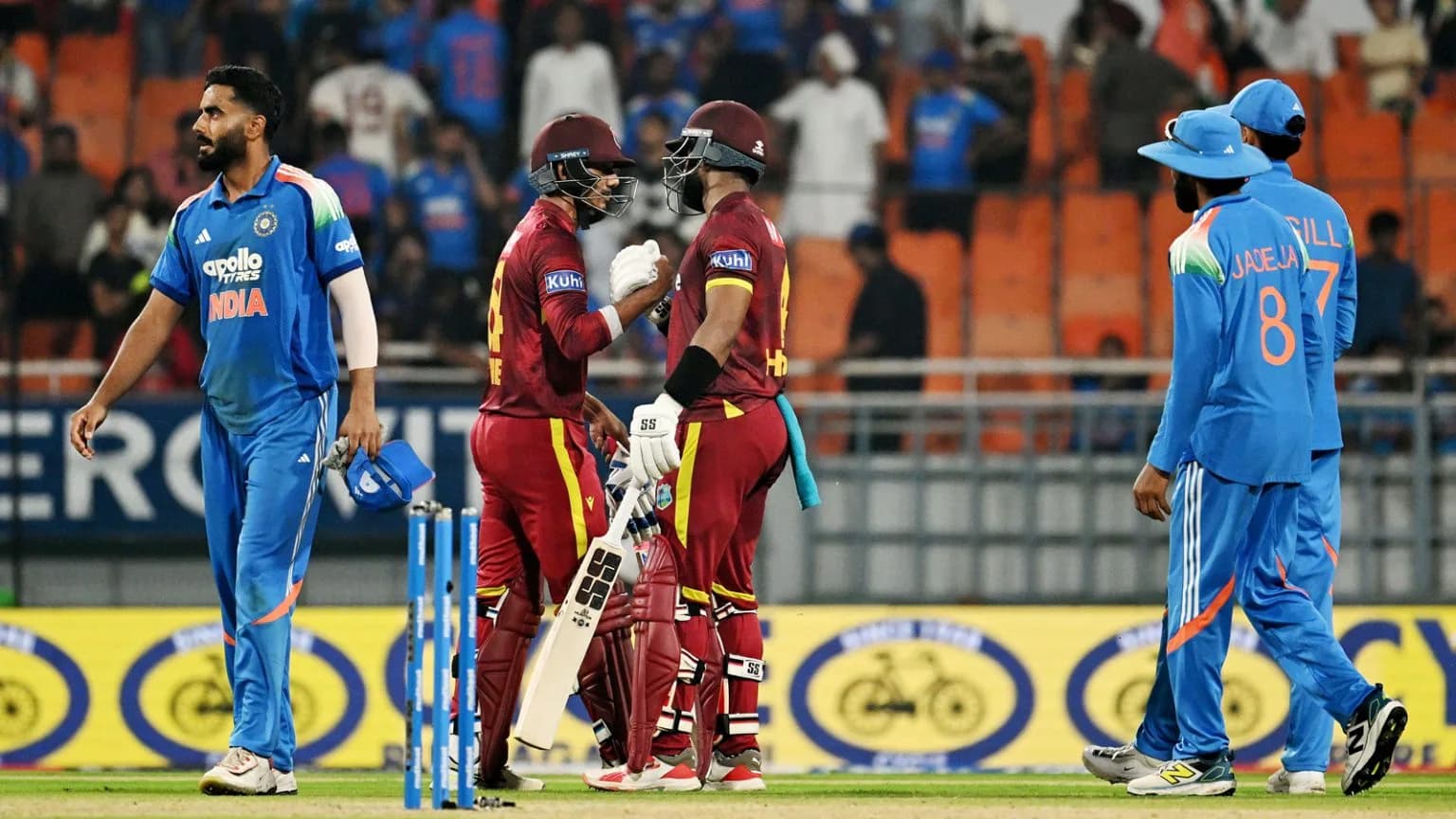 Two West Indies batsmen fist-bumping on the pitch, with Indian players nearby.