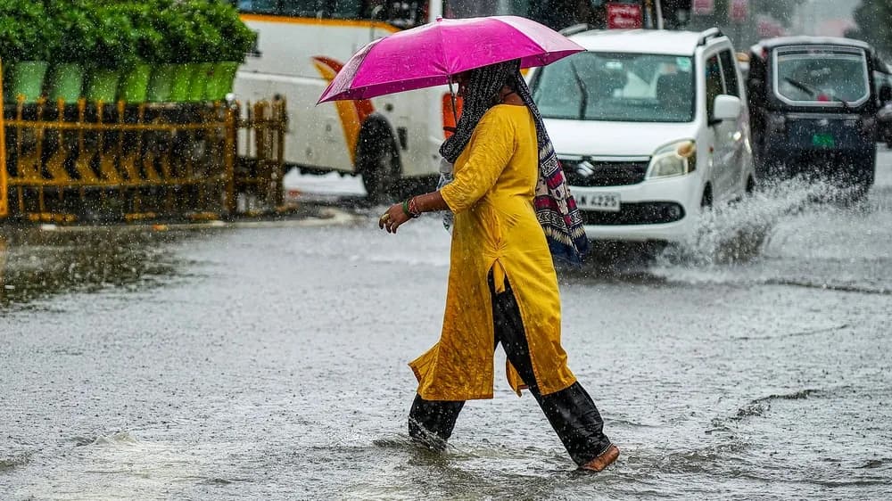 A woman in a yellow dress and black pants walks through a flooded street with a pink umbrella.