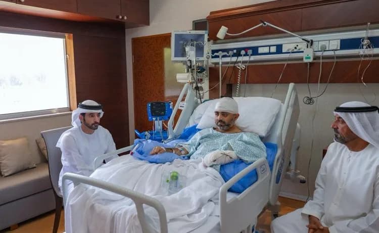 Two men in traditional Emirati clothing visit a patient in a hospital bed.