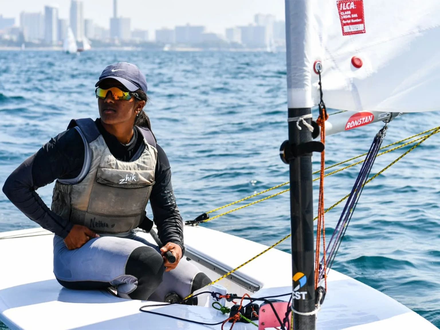 A woman sails a dinghy with a city skyline in the background.