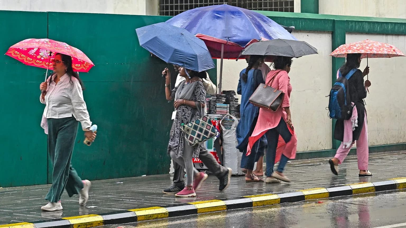 People walk on a wet sidewalk, holding umbrellas in the rain.